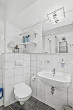 Weiß gefliestes Badezimmer mit Duschkabine, Waschtisch, Schminkspiegel und Pflanzen auf Wandregal. Weiß gefliestes Badezimmer mit Duschkabine, Waschtisch, Schminkspiegel und Pflanzen auf Wandregal.