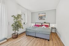 Doppelbett mit grauer Bettwäsche, große Zimmerpflanze, heller Holzboden, schlichte Wanddeko. Doppelbett mit grauer Bettwäsche, große Zimmerpflanze, heller Holzboden, schlichte Wanddeko.