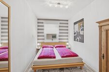 Doppelbett mit pinken Kissen auf Holzrahmen, grau-weiß gestreifte Wandtapete, dezente Deckenbeleucht Doppelbett mit pinken Kissen auf Holzrahmen, grau-weiß gestreifte Wandtapete, dezente Deckenbeleucht