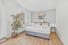 Doppelbett mit weißer Bettwäsche, große Zimmerpflanze, heller Holzboden, schlichte Wanddeko.